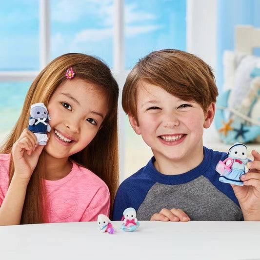 Smiling girl and boy holding Sylvanian Families Seal Family parent figures, with the two miniature baby seals displayed on the table between them.