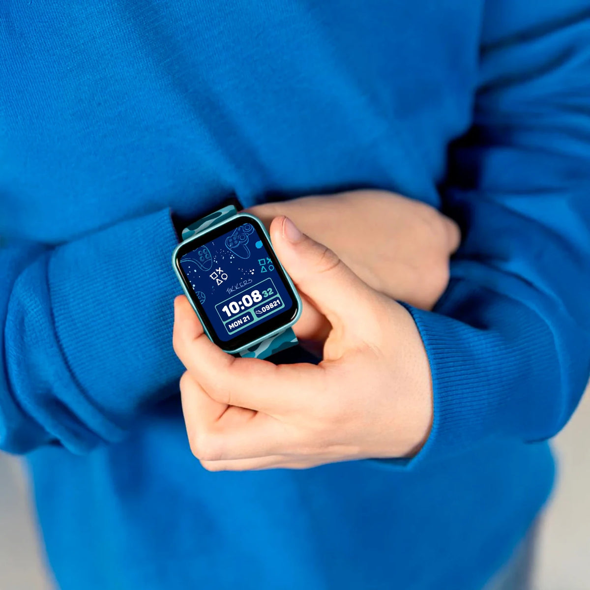 Close-up of a person wearing a Tikkers smartwatch with turquoise case and blue weather display, holding the watch while wearing a bright blue sweatshirt.