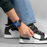 Tikkers Series 13 smartwatch with black silicone strap worn on a wrist beside black and white trainers, displaying a blue digital watch face.