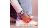 Close-up of a smartwatch with pink digital watch face worn on a wrist beside white trainers with rainbow coloured soles and blue jeans.