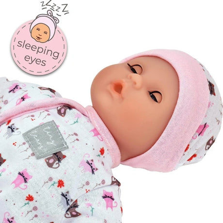 Close view of the Tiny Tears Teeny Baby lying down with closed sleeping eyes, pink trimmed hat and cat print outfit, with a sleeping eyes feature graphic