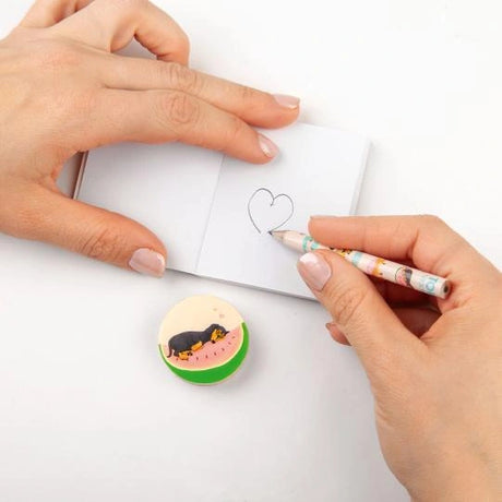 Hands drawing a heart in a small notebook with a TOPModel pencil and round illustrated accessory