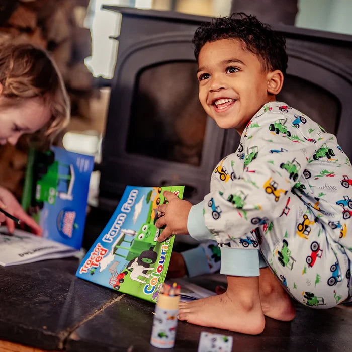 Child holding Tractor Ted Farm Fun Colouring Book while sitting indoors, wearing tractor print pyjamas