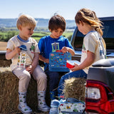 Three children sitting outdoors beside a pickup truck, with one holding the blue Tractor Ted Farmyard Lunch Box decorated with tractors and farm animals.