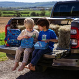 Two children sitting on the tailgate of a pickup truck, opening blue Tractor Ted Farmyard lunchboxes beside hay bales in a countryside setting.