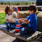 Two children sitting on a pickup truck tailgate beside hay bales, holding Tractor Ted Farmyard Snack Pots with additional containers stacked between them.