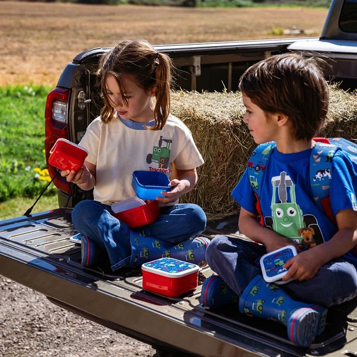 Two children sitting outdoors on a pickup truck tailgate, examining open red and blue Tractor Ted Farmyard Snack Pots with matching illustrated lids.