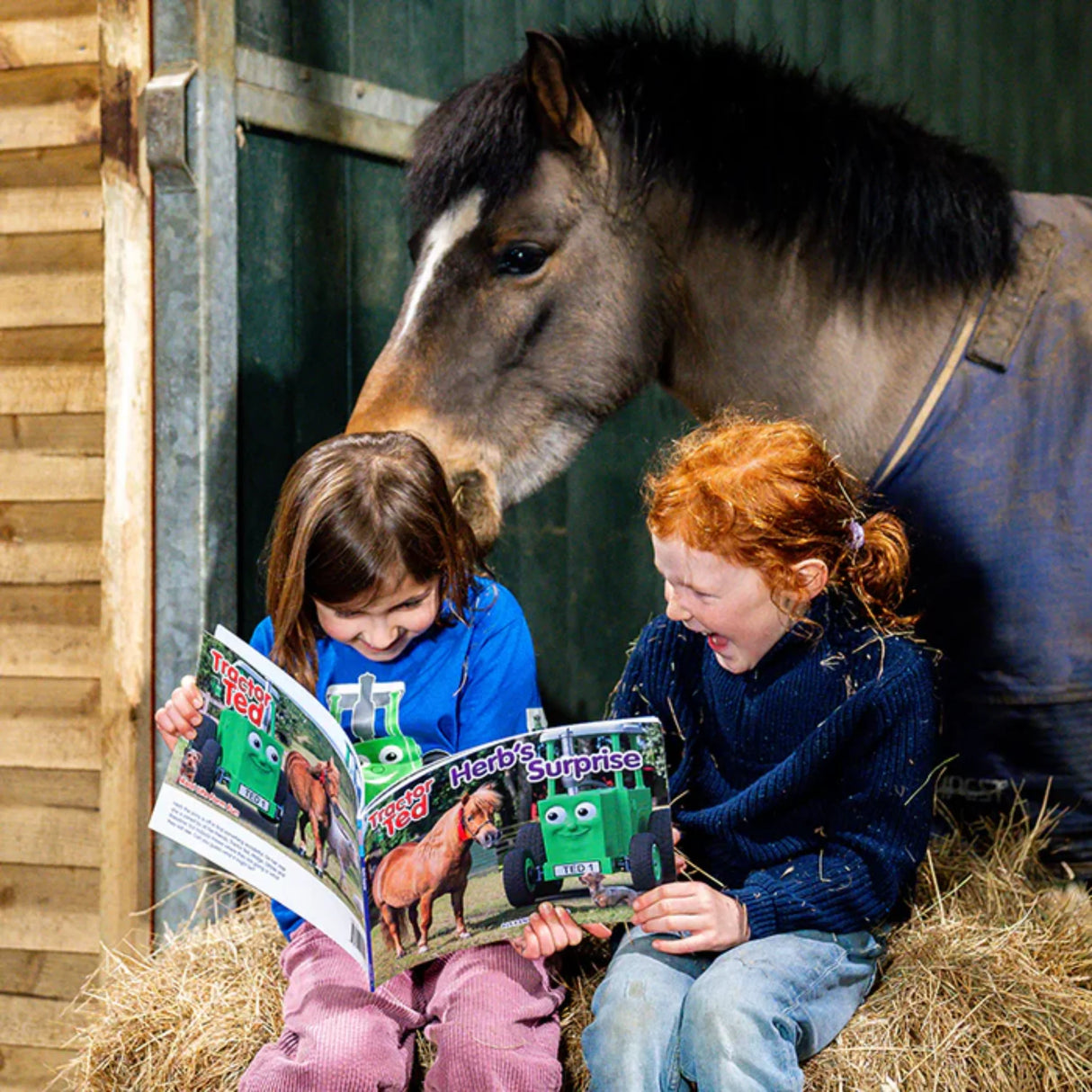 Two children sitting on hay bales reading Tractor Ted Herb’s Surprise Storybook beside a pony in a stable yard