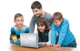 Four children gathered around a VTech Genio MAX laptop on a table, showing the silver back of the screen and wired mouse