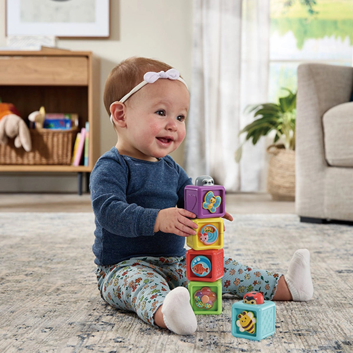 Baby wearing a blue top and patterned trousers sitting on a rug and stacking VTech Pop-a-Blocks, with a bee block beside her