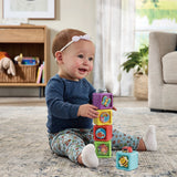 Baby wearing a blue top and patterned trousers sitting on a rug and stacking VTech Pop-a-Blocks, with a bee block beside her