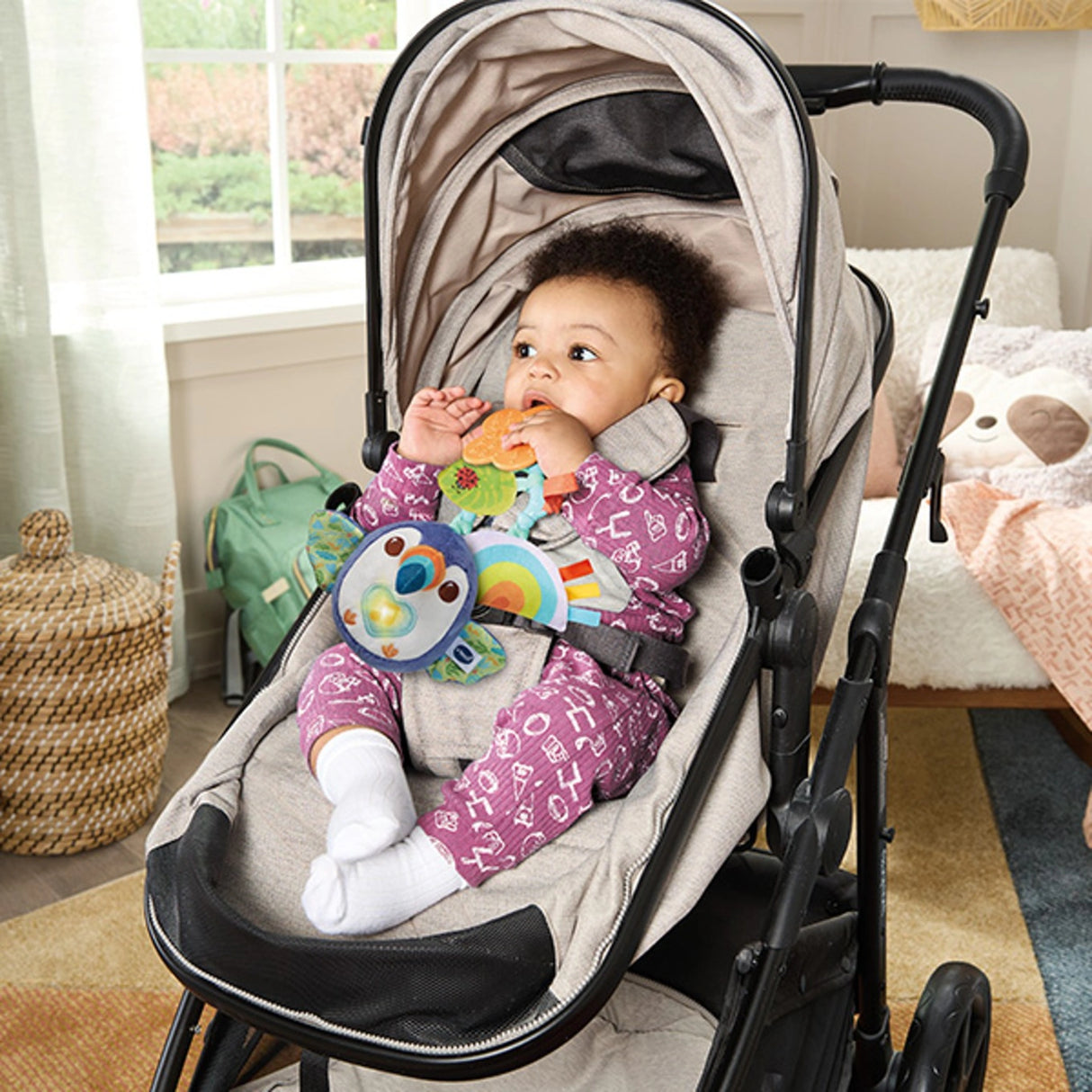 Baby sitting in stroller holding VTech Take Along Toucan toy attached by strap, showing light up heart and rainbow fabric rattle