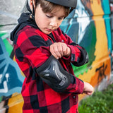 Boy wearing Xootz Kids Pad Set black large adjusting elbow pad while wearing a red check shirt and helmet