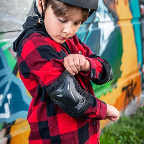Boy wearing Xootz Kids Pad Set black large adjusting elbow pad while wearing a red check shirt and helmet